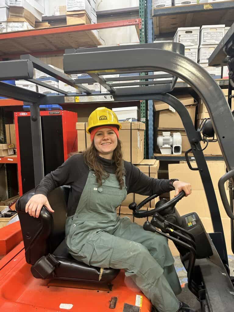 Female worker in safety gear operating forklift in warehouse.