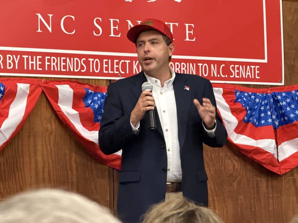 Corbin speaking at campaign event with patriotic decorations in background.