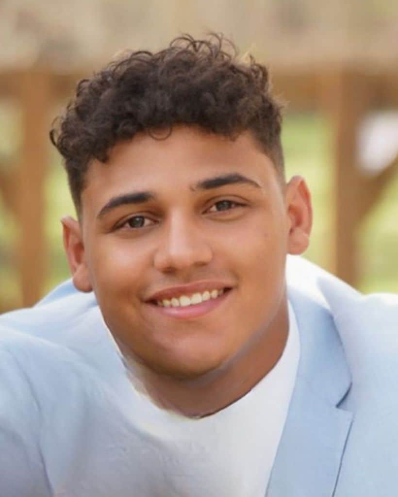Smiling young man with curly hair in a light blue blazer outdoors.