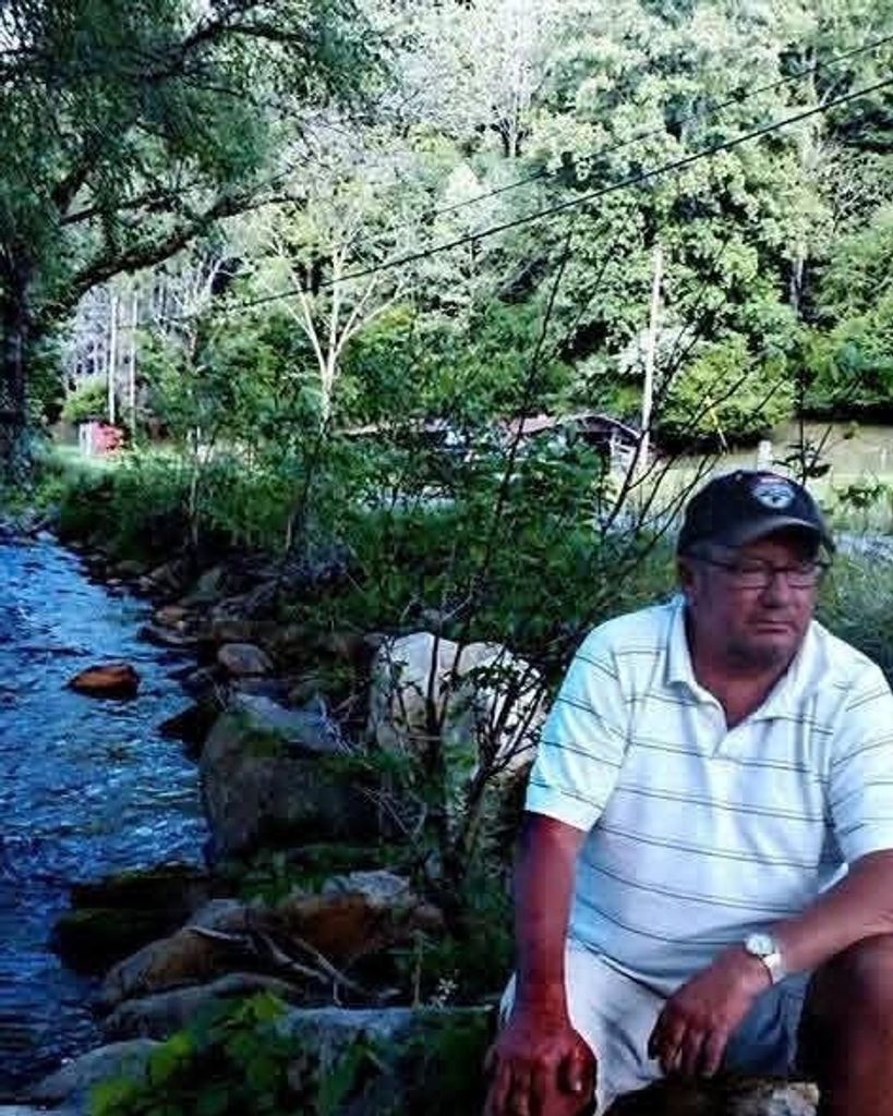 Man sitting by a creek with trees and water in the background.