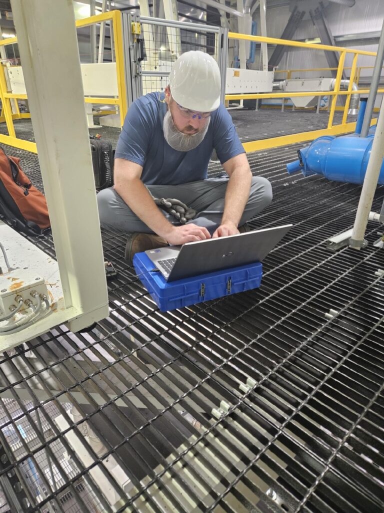Engineer working on industrial equipment with laptop in factory setting.