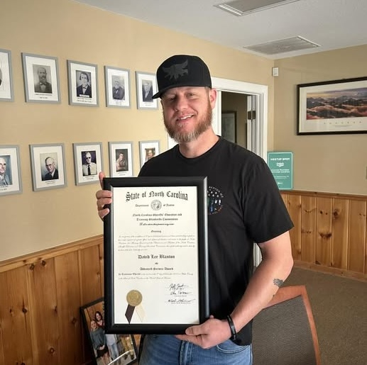 Man holding a framed certificate in an office setting with framed photos on the wall.
