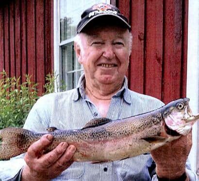Elderly man displaying a large fish outdoors in front of a red wooden building.
