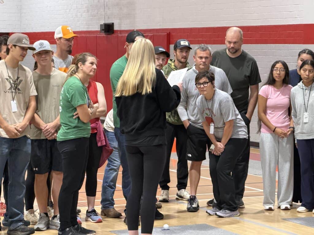 Group of people listening to a woman speaking in a gymnasium.