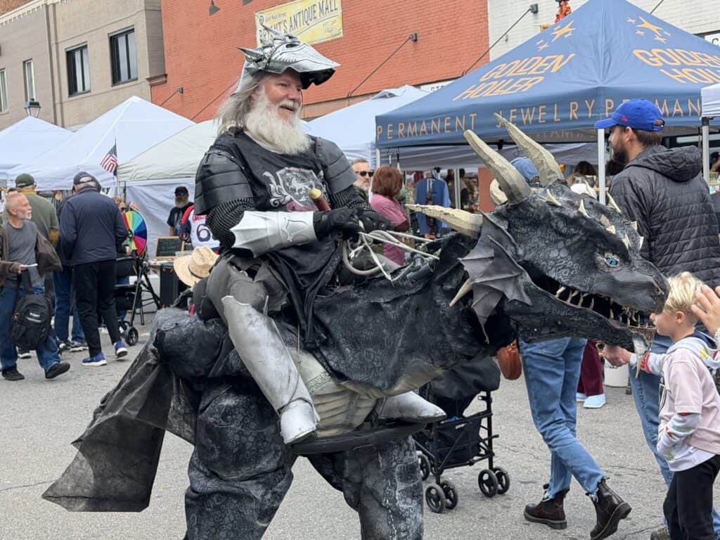 Person in dragon costume at fall festival with children and tents.