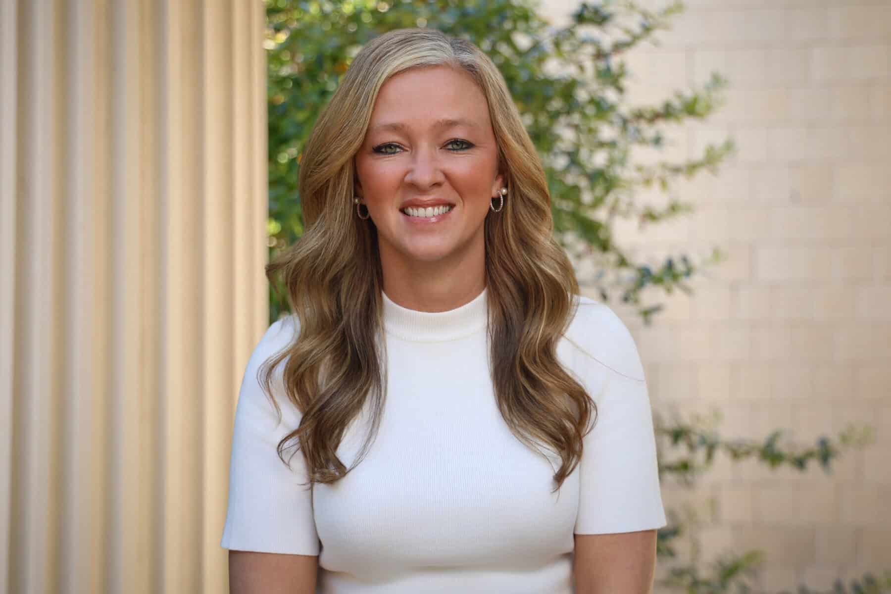 Smiling woman with long wavy hair in white top outdoors.