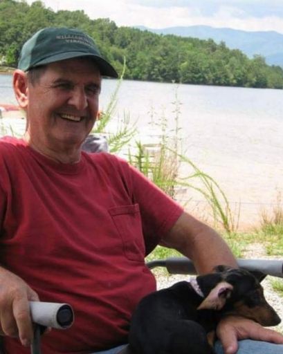 Man smiling with dog near lake in outdoor setting.